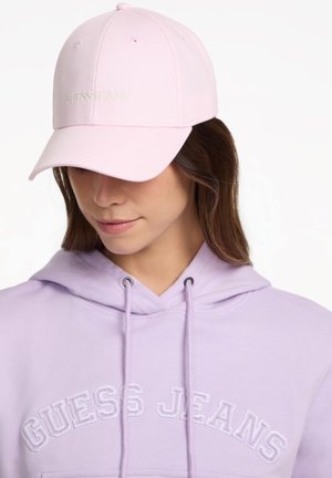 Femme portant une casquette Guess Jeans rose clair et un sweat à capuche Guess Jeans lavande, regardant vers le bas avec des cheveux partiellement visibles.