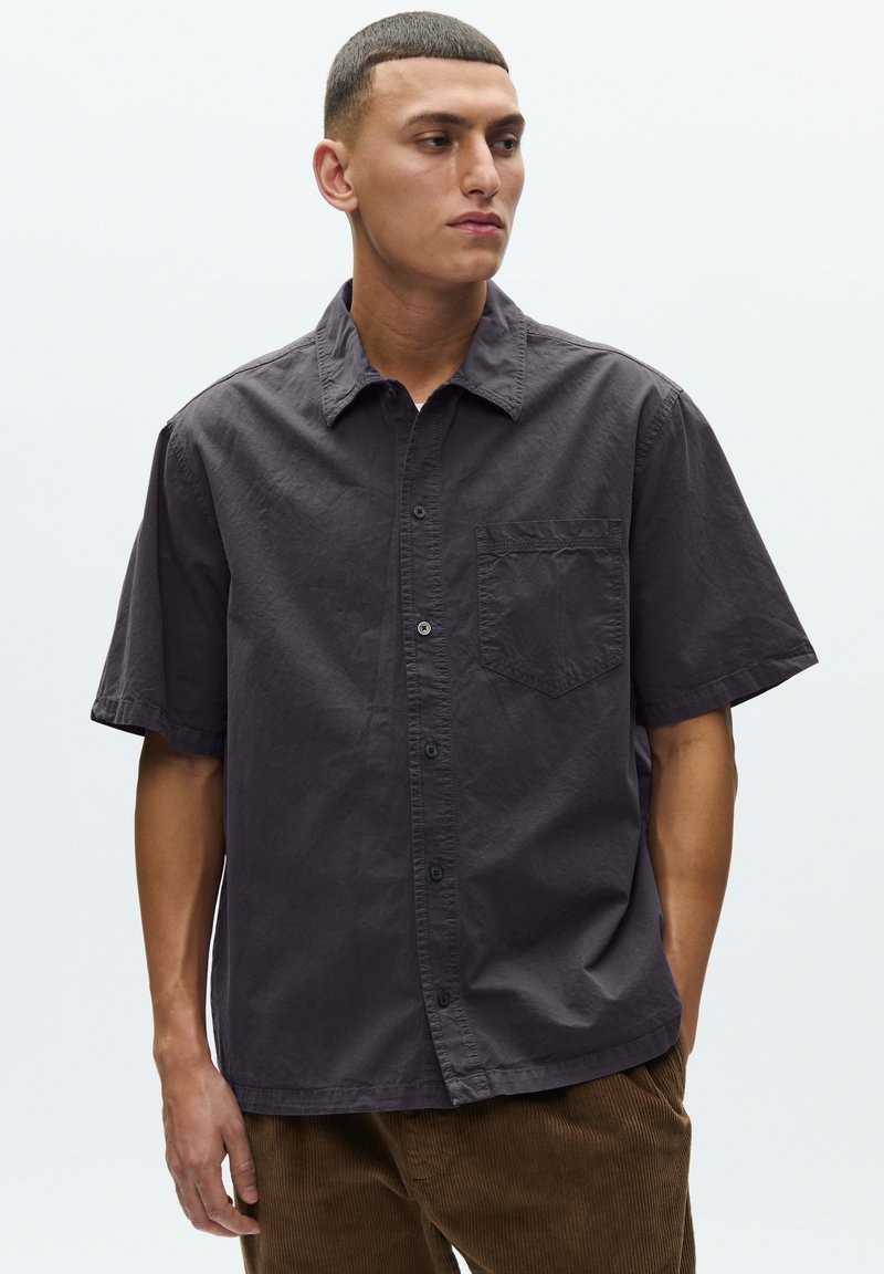 Noormees, kes kannab lühikeste varrukatega tumedat halli nööbitavat särk ja pruune pükse, vaatab veidi vasakule, taust on lihtne.