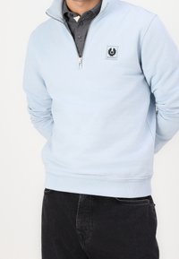 Uomo che indossa una felpa a zip metà con un colore blu chiaro sopra una polo grigia scura, abbinata a jeans neri.