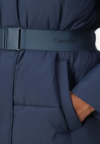 Tamsiai mėlynas pūkų paltas su lygia tekstūra, turintis platų diržą su "Calvin Klein" logotipu ir šoninėmis kišenėmis.