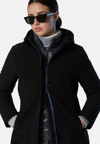 Giacca esterna nera con zip frontale, abbinata a una giacca interna trapuntata. Dolcevita grigio e polsini testurizzati. Occhiali da sole scuri.