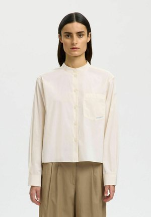 Femme aux cheveux lisses et foncés, portant une chemise crème boutonnée avec une poche poitrine et un pantalon ample beige, debout devant un fond uni.