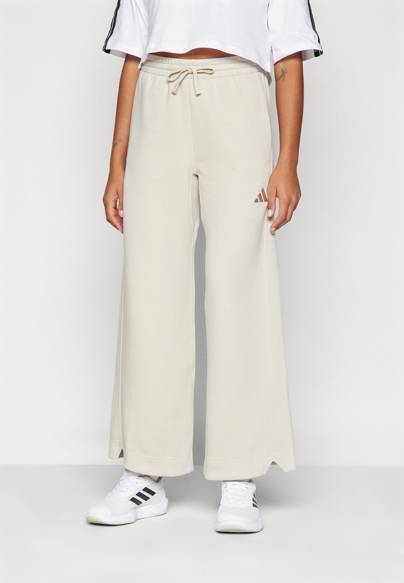 Beige széles lábú melegítőnadrág rugalmas derékkal és húzózsinórral, a bal combon egy kis barna Adidas logóval.