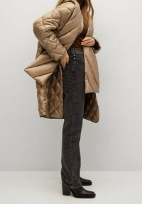 Cappotto beige trapuntato con finitura satinata, abbinato a jeans a vita alta grigio scuro. Stivali neri con tacco completano l'outfit.
