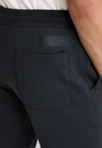 Dunkelblaue Jogginghose mit glatter Textur, ausgestattet mit einer Gesäßtasche und einem gestickten Logoetikett am Bund.