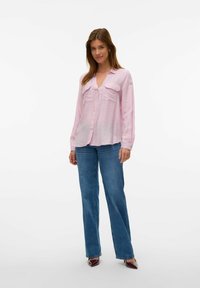 Camisa de botones de color rosa claro con dos bolsillos en el pecho, mangas largas y un corte relajado, combinada con unos jeans rectos de color azul oscuro.
