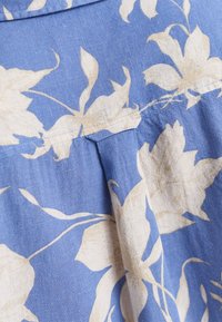 Blauer Stoff mit einem floralem cremefarbenen Muster, der eine strukturierte Oberfläche und ein plissiertes Design mit sichtbaren Nähdetails aufweist.