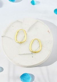 Guldtonade hoopsmycken med en förlängd tårform, visade på en strukturerad betongplatta med spridda blå glaselement.