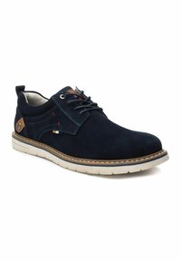 Zapato de ante azul marino con una textura suave, acentos de cuero marrón en contraste y una suela de goma blanca. Presenta cierre con cordones y costuras decorativas.