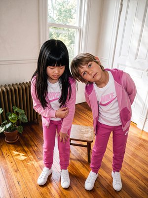Deux filles vêtues de survêtements roses assortis se penchent vers l’appareil photo dans une pièce lumineuse au sol en bois.