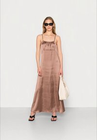 Vestido de cetim bronze com alças finas, forma A-line e decote franzido. Usado com sandálias pretas e a segurar uma bolsa de ombro leve.