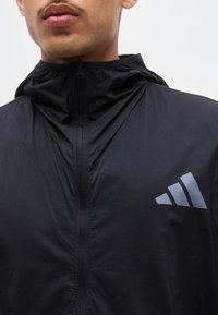 Homme portant une veste noire à capuche zippée avec un logo Adidas gris sur la poitrine gauche, montré du cou jusqu'à la moitié du torse.