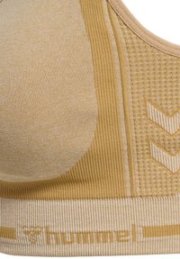 Nahaufnahme eines senfgelben, gerippten Sport-BHs mit strukturierten Mustern und dem "hummel"-Logo am unteren Bund.