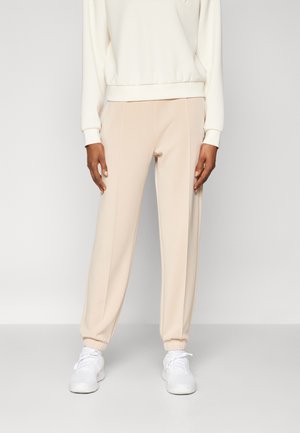 Person trägt beigefarbene, verjüngte Hosen mit elastischen Bündchen, weiße Sneakers und ein elfenbeinfarbenes langärmeliges Oberteil und steht vor einem einfarbigen Hintergrund.