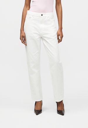 Žena oblečená v bielych nohaviciach s vysokým pásom a rovnými nohavicami, svetloružovom priliehavom topu a čiernych lodičkách s ostrou špičkou, stojaca pred jednofarebným pozadím.