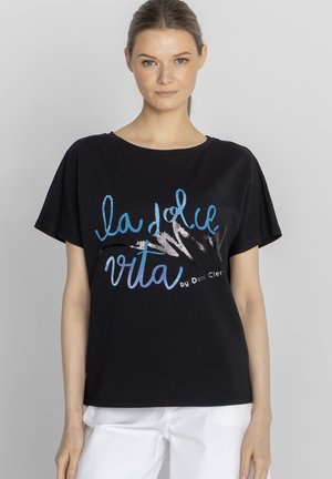 Kobieta w czarnym T-shircie z niebieskim i białym napisem „la dolce vita by Deni Clèr” oraz w białych spodniach na jednolitym tle.