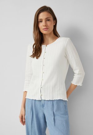 Jeune femme aux cheveux bruns porte un cardigan en maille blanc boutonné avec des manches trois-quarts et un jean bleu clair ample, debout contre un mur gris.