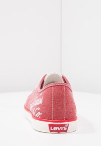 Červená plátěná teniska s bílým šitím, logo "Levi's" a kontrastní gumová podrážka. Nízký profil a zaoblený tvar. Styl na nazouvání.