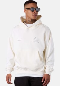 Cream-colored hoodie with a front pouch pocket, ribbed cuffs, and hem. Features printed logo on the left chest. Soft fabric texture.