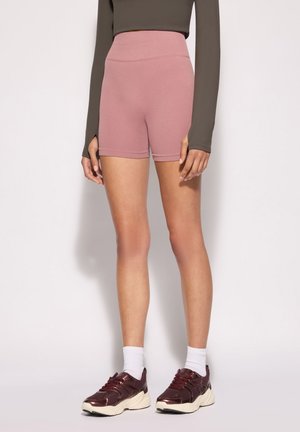 Persona che indossa pantaloncini rosa a vita alta, top oliva a maniche lunghe con fori per i pollici, calzini bianchi e sneakers bordeaux, in piedi su uno sfondo semplice.
