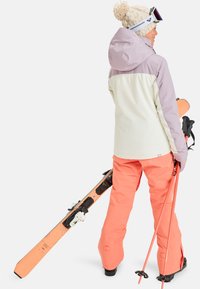 Ski outfit features a dual-toned jacket with a light purple and cream color scheme, paired with bright orange ski pants, holding skis and poles.