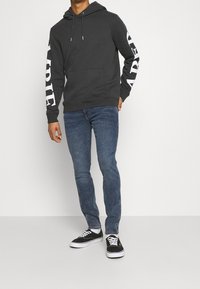 Tmavě šedá mikina s kapucí a přední kapsou, s bílým textem na rukávech, v kombinaci s modrými džíny a černými teniskami. Jednoduchý, ležérní design.