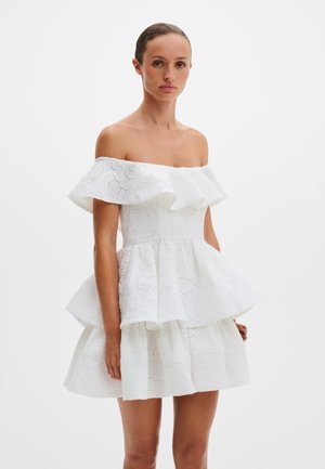 Donna che indossa un vestito bianco a strati con spalle scoperte, ricamato a motivi floreali traforati e dettagli arricciati, in piedi davanti a uno sfondo semplice.