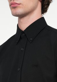 Μαύρο πουκάμισο με κουμπιά, με δομημένο γιακά, απαλό υφή και διακριτική κεντημένη λογότυπο στο στήθο�ς. Στρογγυλά μαύρα κουμπιά διακοσμούν το μπροστινό μέρος.