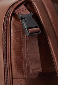 Détail d'un sac en cuir marron montrant une boucle en plastique noire et une sangle en tissu texturé, mettant en évidence les coutures et le contour de la fermeture éclair.