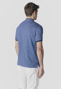 Homme de dos portant un polo bleu à manches courtes et un short cargo blanc, sur un fond clair uni.