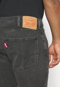 Poche arrière d'un jean Levi's noir avec une étiquette rouge Levi's et un patch en cuir beige de la marque sur la ceinture, porté par une personne avec un bras droit visible.