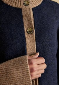 Zapínanie gombíka na námorní modrom a pieskovom pletenom kardigáne s rebrovaným golierom a manžetami.