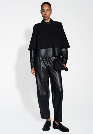 Zwarte gebreide cape met een brede silhoutte, gecombineerd met zwarte leren broeken. De outfit heeft een geknoopte kraag en een gestructureerde hoed.