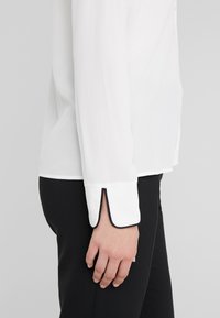 Witte blouse met lange mouwen, subtiele glans en zwarte bies langs de manchetten. Glad, lichtgewicht stof, ingestopt in zwarte broek.