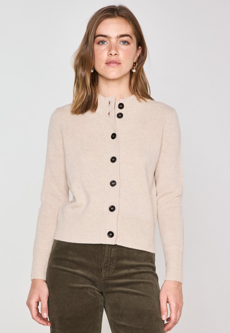 Cardigan beige clair en matière douce, avec un col rond et sept boutons noirs sur le devant. Bords ribbés aux poignets et à la taille.