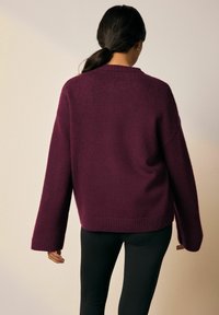 Nainen, jolla on tumma poninhäntä, pukeutuneena löysään, pitkähihaiseen, tummanpunaiseen neuleeseen ja mustiin housuihin, kääntyy poispäin kamerasta.