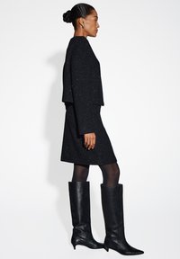 Schwarzes Tweedkleid mit metallischen Einsprengseln, strukturierte kurze Jacke. Model trägt knielange schwarze Lederstiefel mit spitzen Zehen. Glatte Textur.