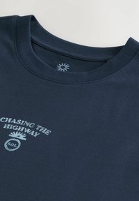 T-shirt bleu foncé à col rond avec texte bleu clair "CHASING THE HIGHWAY" et un petit emblème circulaire sous le texte, près de l'encolure.