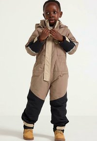 Skidragt i beige og sort med vandtæt ydermateriale, hætte, reflekterende detaljer og elastiske manchetter. Beige støvler fuldender outfittet.