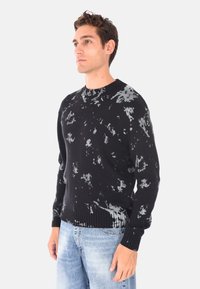 Maglione nero a maglia con un motivo distressed grigio, collo rotondo e polsini e orlo a costine. Indossato con jeans azzurri chiari.