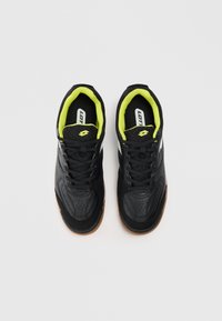 Um par de sapatilhas pretas de desporto com forro interior amarelo-neon e solas castanhas, vistas de cima sobre um fundo branco.