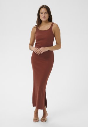 Kobieta o długich brązowych włosach, ubrana w bezrękawnik, rdzawobrązową sukienkę sięgającą do kostek oraz beżowe buty na obcasie z odkrytymi palcami, stojąca z rękami złożonymi razem.