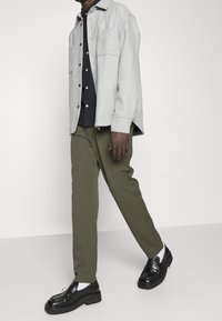 Graues Wolle-Mix-Hemdjackett über einem dunkelblauen Button-Up, kombiniert mit olivgrünen Hosen und schwarzen Slippern. Weiße Socken sind sichtbar.