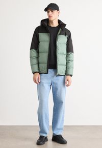Gevoerde groene en zwarte jas met een capuchon, gewatteerde textuur, ritssluiting en verborgen zakken, gecombineerd met lichtblauwe jeans en zwarte schoenen.