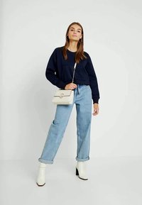 Sweatshirt cropped azul-marinho, jeans de cintura alta azul-claro, botas de tornozelo brancas, e uma bolsa creme com um detalhe circular.