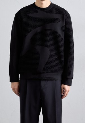 Homme portant un sweat-shirt noir avec un grand motif graphique tonal et un motif de texte, associé à un pantalon noir à jambes larges, debout devant un fond uni.