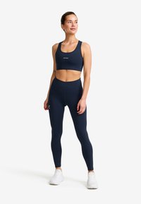 Kvinna som står, klädd i marinblå sportbh och leggings med vita sneakers, ser lätt åt höger, med håret uppsatt.