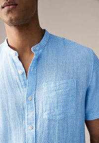 Hele sinine lühikeste varrukatega särk tekstuurse koega, millel on püstkrae, rinnatasku ja valged nupud.