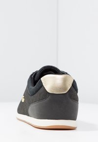 Svart sneaker med vit sula och guldhäll, visad bakifrån på en vit yta med en plain ljusgrå bakgrund.