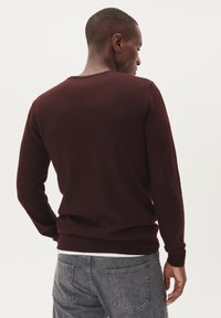 Mörkröd stickad tröja med långa ärmar och rundad halsringning. Har en ribbad nederkant och ärmslut, bärs över en vit skjorta, med en slät struktur.